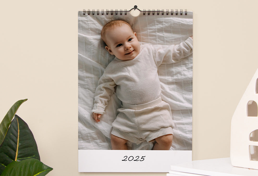 Calendrier murale A3 portrait accroché à un mur et ouvert sur la page de couverture qui montre une grande photo d'un bébé avec le texte "2025" dessous.