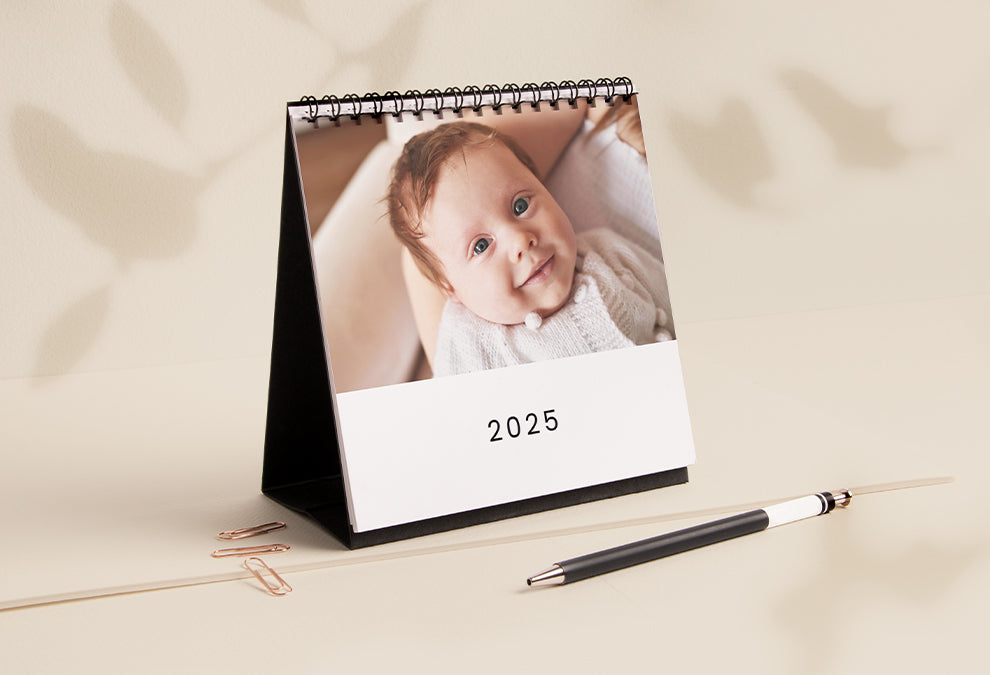 Calendrier de bureau au format carré posé sur une table ouvert à la page de couverture avec la photo d'un bébé et le texte "2025" en dessous.