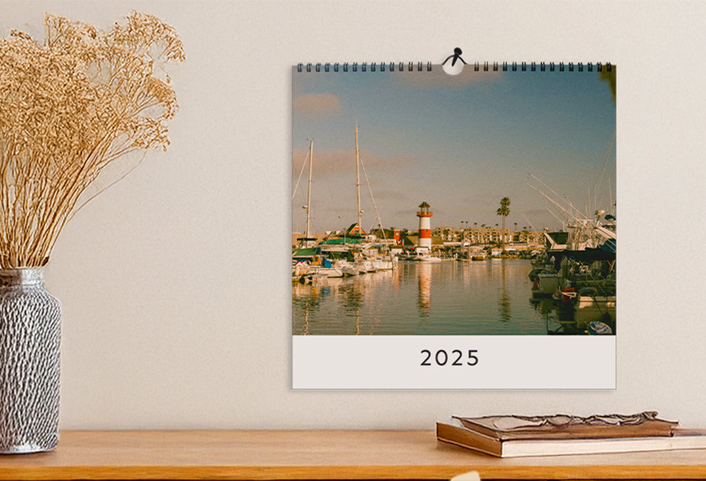 Calendrier murale carré accroché à un mur et ouvert sur la page de couverture qui montre une grande photo d'un port et le texte "2025" dessous.
